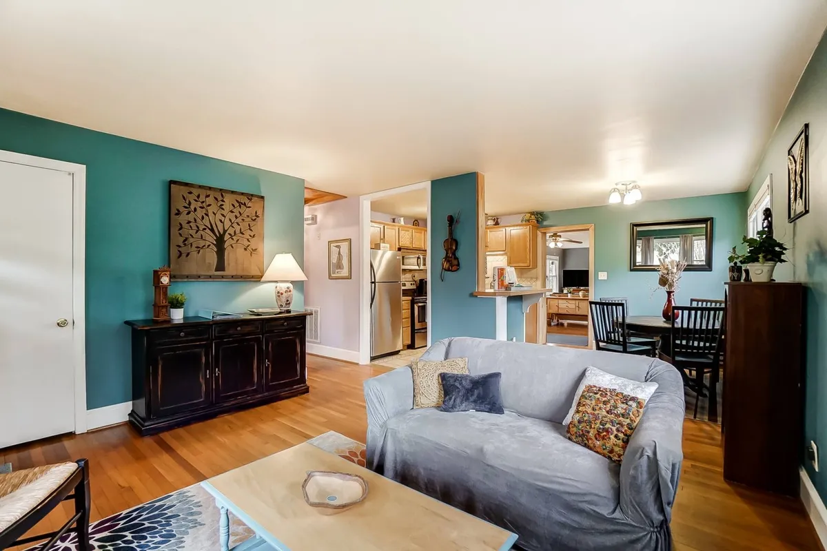 This image showcases a cozy living area with a sofa decorated with vibrant pillows, stylish wall art, and a black cabinet. The open layout reveals a kitchen area in the background and a dining table, suggesting a versatile space ideal for gatherings. The decor gives off a bohemian flair, inviting relaxation and social activities.