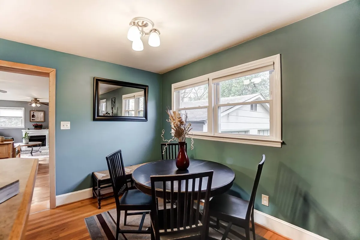 A cozy dining area with a round table, black wooden chairs, a vase with dried flowers, and a mirror on the wall. The setting is intimate, perfect for quick breakfasts, family meals, or late-night chats, and is warmly lit, making it feel inviting and versatile.