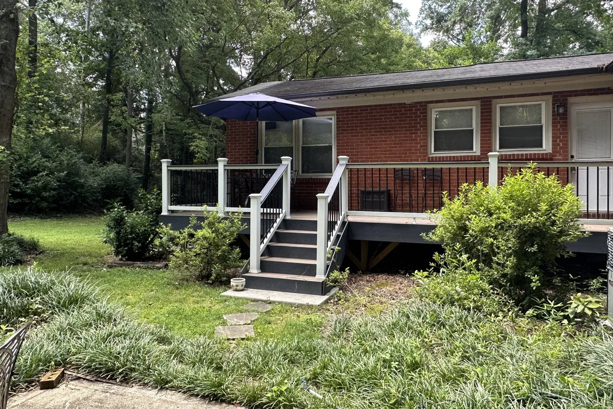A welcoming exterior view of the home's backyard area, featuring a wooden deck with steps leading to the door, shaded by an umbrella, and surrounded by lush greenery. The partially fenced yard is visible, creating a sense of a secure, pet-friendly outdoor space.