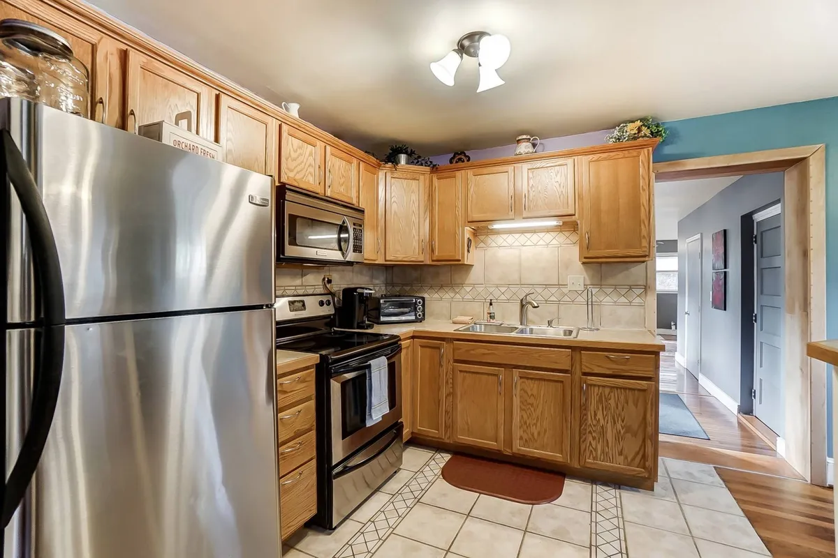 A close-up view of a kitchen area with stainless steel appliances, a kitchen sink, wooden cabinetry, and a tile backsplash. The scene feels homey and functional, reflecting a space where daily routines like coffee-making and cooking unfold.