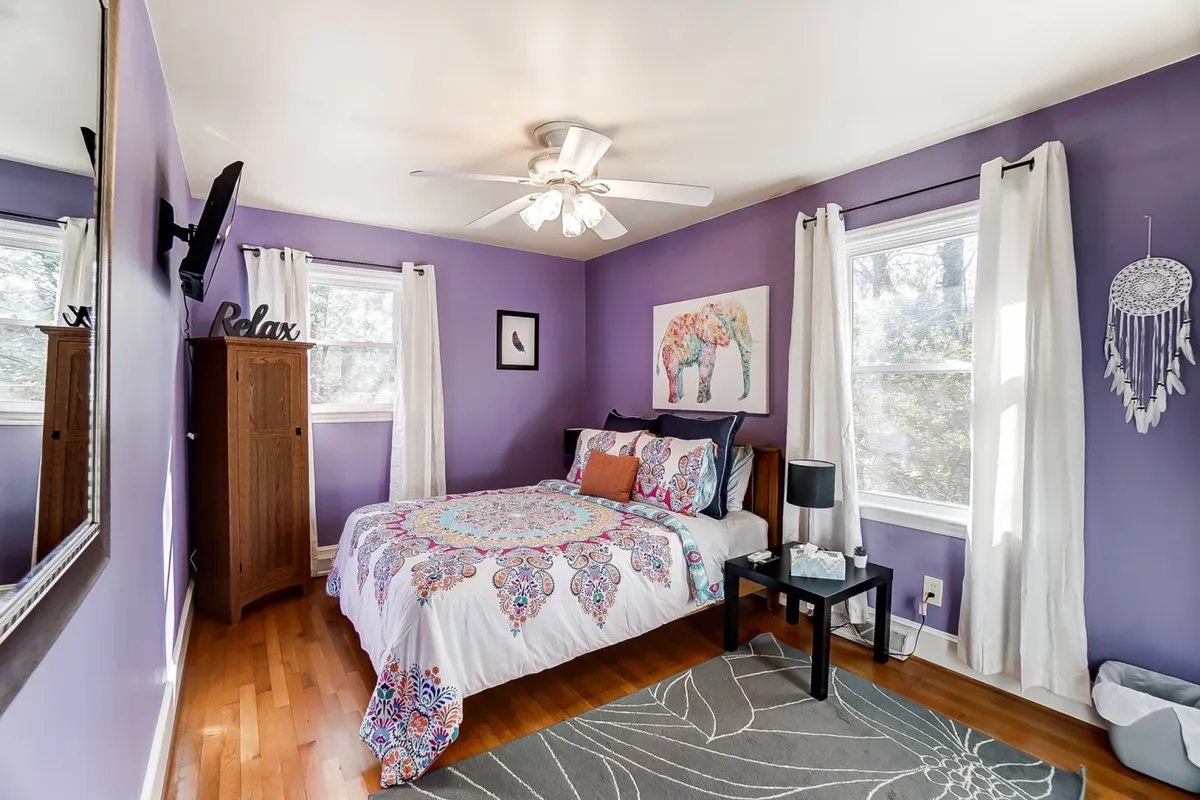 A cozy bedroom with a colorful bedspread, elephant artwork above the bed, a wooden wardrobe, a wall mirror, and a soft floor rug. The room is warmly lit and inviting, with sunlight streaming in through a window.