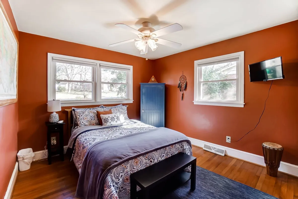 A welcoming bedroom with decorative bedding on a bed, two windows allowing abundant natural light, a blue wardrobe, and a small television mounted on the wall. The setting is clean, comfortable, and homey.