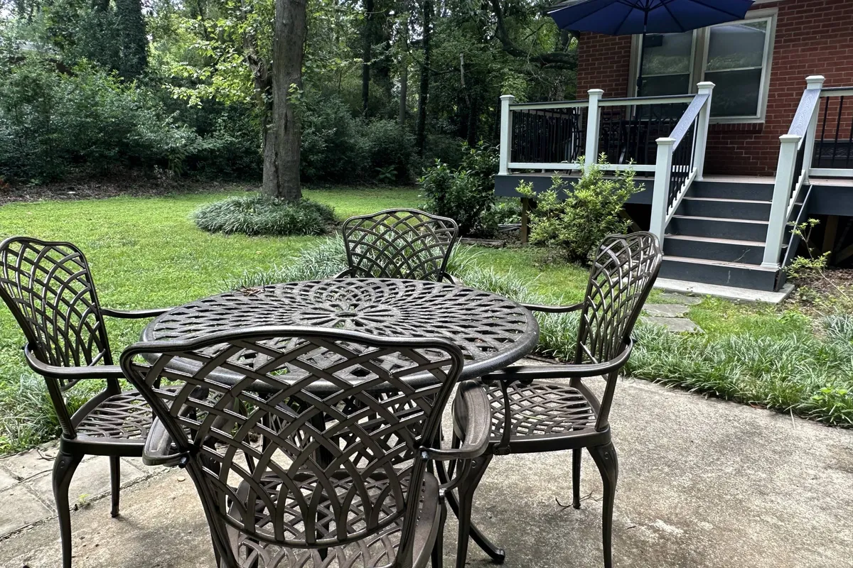 A cozy exterior scene featuring a decorative metal table and woven chairs set on a green lawn. The space is adjacent to a brick house with steps leading up, and a large blue umbrella provides shade. The area feels welcoming and is suitable for both human and pet relaxation or play, with enough room for pets to enjoy the outdoors.